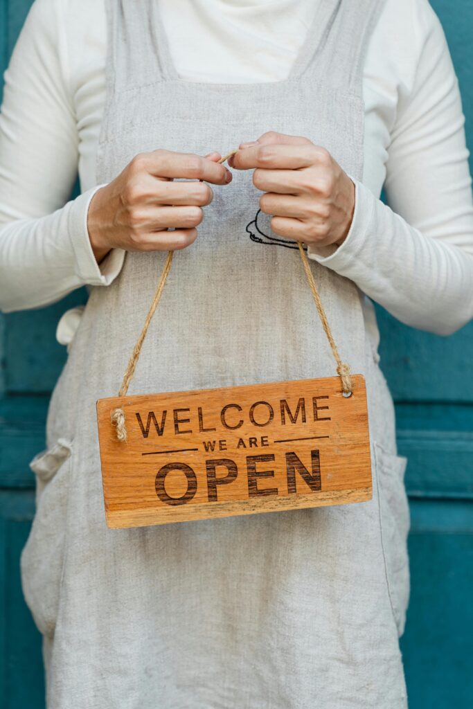 pexels-photo-4473360-4473360 Crop faceless female in casual clothes and apron demonstrating wooden sign with text welcome we are open against blue door